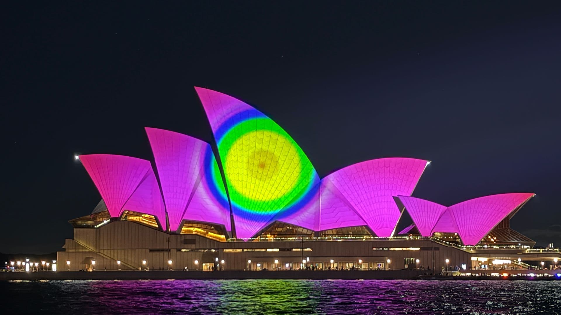 Sydney Opera House illuminated in pink with blue, green and yellow circular light projection during Vivid Festival, reflections over Circular Quay