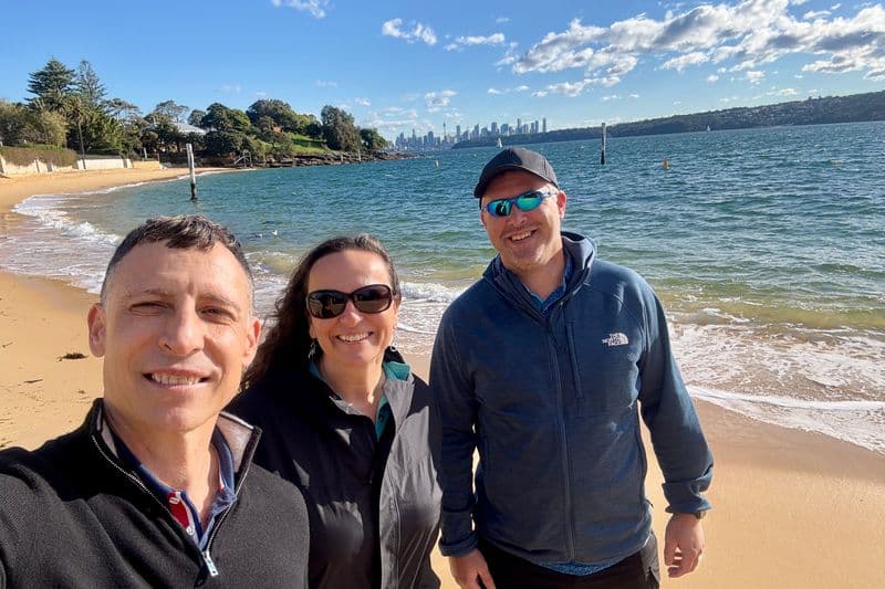 Tour guide and couple at Camp Cove beach, Sydney Harbour, with city skyline visible across calm harbour waters.