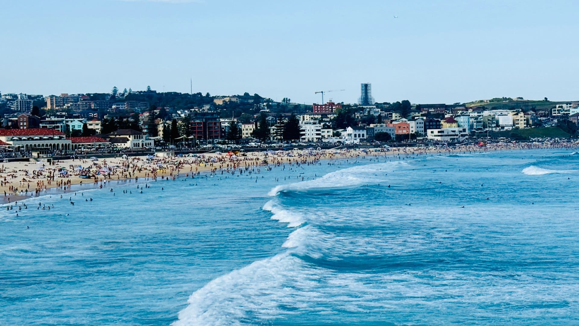 Bondi Beach on a busy sunny day — crowds on golden sand as a long white wave rolls toward swimmers in the shallows.